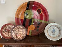 Four decorative plates displayed with individual stands, including a large abstract wooden plate, two smaller plates with bird designs, and a white plate with a religious figure.