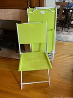 Two bright lime green folding chairs with polyester mesh fabric on white metal frames, one chair open and one folded, shown indoors on a wooden floor.