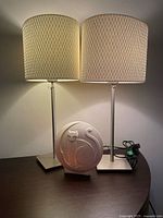 Two height adjustable table lamps with brushed metal square bases and beige textured fabric cylinder lampshades, alongside a white ceramic decorative cat nightlight on a dark wood surface.