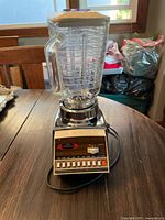Vintage kitchen blender with clear plastic blending pitcher, multiple control buttons on base, chrome and beige design, placed on wooden table.