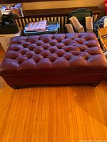 Full view of the rectangular burgundy faux leather storage ottoman on wooden floor in front of wooden furniture, showing tufted button top and overall structure.