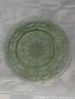 Top-down view of 10 inch uranium Vaseline glass plate on white textured cloth backdrop, showing central geometric and floral pattern in translucent green color.