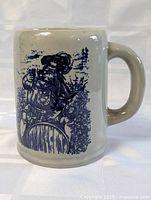 Front view of antique stoneware mug showing detailed blue illustration of a man holding a shield with plant details on off-white stoneware.