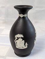 Front and side views showing black bud vase with white classical relief cherub and foliage decorations on matte surface.