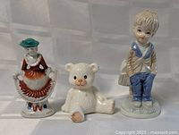 Three ceramic figurines: woman in bonnet and dress, teddy bear, boy with sack, arranged side by side