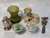 Green olive glass pitcher, small bear figurine with honey pot, metal shaker, two floral ceramic candle holders, and moose-themed spreader arranged on a white surface.