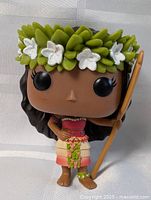 Front view of Hana Lolo Funko Pop figure showing detailed facial features, leaf and flower headband, red top, grass skirt with flower accents, and brown spear held in right hand.