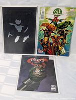 Photo showing three comics: black cover with shadow figure, colorful Marvel Avengers comic, and comic titled 1602.