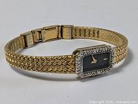 Gold filled woven bracelet watch with rectangular black dial and CZ stone bezel, lying flat showing bracelet and clasp.