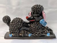 Front view of black ceramic poodle with curled fur texture and pink ears, red nose, blue bow, blue paws laying on rectangular base.