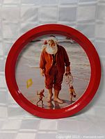 Round metal tin with a red rim and vintage style image of barefoot Santa Claus at beach alongside elves and kite.
