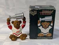 Teddy bear figurine sitting holding silver Stanley Cup trophy overhead, next to original box showing the front panel with matching design.