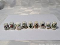 Front view of the full set of 9 painted porcelain thimbles with various floral and nature themes arranged in a row on a white background