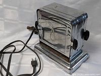 Front and side view of vintage stainless steel chrome toaster showing adjustment knobs, electric cord and reflective metal surface.