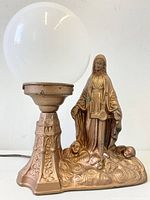 View of entire lamp showing detailed metal base with embossed designs, Mother Mary statue standing on cloud base with cherubs, and large white glass globe light on side.