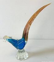 Side angle showing vibrant blue body and long amber tail of Murano art glass pheasant bird on a clear scalloped glass base.