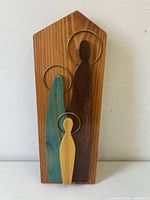 Front view of the carved wood Holy Family wall sculpture showing the three abstract figures with metal halo rings mounted on a natural-tone wooden backing shaped like a house.