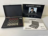 Open electronic chess computer showing built-in sensor chessboard with red and white pieces, alongside three manuals including Kasparov Chess, Cavalier user manual, and Let's Play Chess USCF rules booklet.