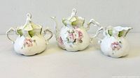 Front view of whole three-piece porcelain set with pink rose designs and green leaf accents, showing teapot, creamer, and sugar bowl.