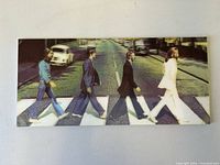 Front view of the rectangular hologram picture on board showing The Beatles Abbey Road crossing with members walking from right to left wearing distinct outfits, white stripes of the zebra crossing, and a white Volkswagen Beetle on street.