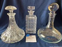 Three clear glass vintage decanters displayed against a dark blue background, showing varying designs and stoppers.