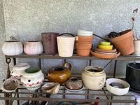Full view of assorted planting pots arranged on metal shelves against a textured wall, including terracotta, ceramic, glazed, jug-shaped pots and abalone shells.