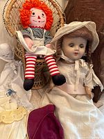 Antique dolls including Raggedy Ann doll, antique doll with braids, ghost-like cloth doll, bonnet, yellow cloth, and wicker doll chair