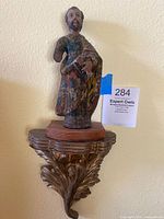 Full view of the painted wooden religious figure mounted on a carved gold-painted wooden base attached to a wall.