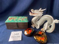 Photo showing white ceramic dragon figurine next to cloth covered embroidered jewelry box and two black lacquered stones with fish design