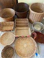 Full view of the collection showing various shaped woven baskets arranged on the floor close together: round, rectangular, cylindrical, some with handles, one with flower decoration