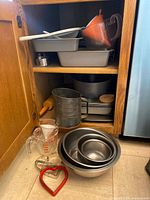 Kitchen shelf showing nested metal mixing bowls, loaf pans, round cake pans, measuring cups, flour sifter, and other baking accessories