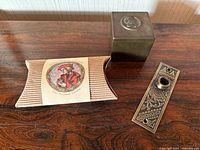 Photo displaying all three items on a wooden surface: the brass square holder, decorative doorbell buzzer frame, and cardboard box with sundial label.