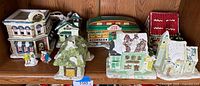 Full display of seven ceramic Christmas houses and two ceramic couples on wood shelf with varied shapes, colors, and lighting cords visible.