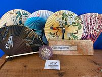 Lot showing five Asian decorative folding fans, a small cocktail umbrella, and the Chinese Buddhist book with fabric cover placed on a wooden surface against a blue background.