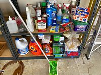 Two shelves filled with various cleaning products including large detergent bottles, boxes of dryer sheets, cleaning sprays, and mop.