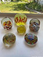 Set of five colorful handblown glass paperweights displayed together on a window ledge with green yard visible in background, showing various shapes and vibrant internal patterns.
