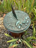 Photo showing copper sundial set in ground with visible Roman numerals, sun motif, and green patina.