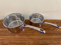 Front angled view of two stainless steel pots with handles, on wood surface against beige wall.