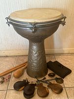 Full view of lot on tile floor showing metal goblet drum with skin head, two wooden drumsticks, castanets, small wooden beater, small cymbal or bell, and black cloth pouch.