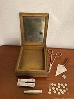 Overview of the wooden antique box with mirror, small scissors, Freemason triangular metal tool with wooden handle, metal Freemason pin, metal square pin, Leatherman multi-tool, small folding knife, and an assortment of miniature coins in front of the box