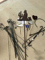 Array of metal garden stakes laid out on a concrete surface, including various decorative tops such as hearts, butterfly, wolf, cactus and angel designs.