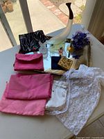 Photo showing a gold toned rectangular tray with various vintage perfume bottles, pink scarves, a white lace blouse, a fabric sachet, a decorative horn or vase, a small wooden box, and a purple faux flower bunch arranged on the tray and table.