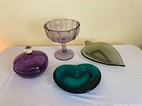 Four glass candy dishes on a white surface against a light yellow wall: purple lidded round dish, purple pedestal bowl, olive green leaf-shaped dish, teal kidney-shaped dish