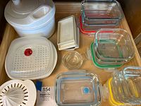 Top-down view of drawer showing glass storage containers with colorful lids, white salad spinner, small glass juicer, ice cube trays, and butter dishes grouped together.