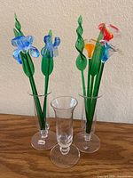 Complete view of art glass flowers arranged in three clear glass vases on wooden surface, showing colorful flower details and green twisted stems.