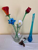 Lot of four hand blown glass flowers displayed in a clear glass vase next to a tall blue glass vase and steel rose sculpture on white cloth.