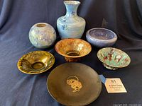 Seven handcrafted ceramic items including vases, bowls, and a decorative plate, shown on dark fabric background.