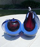 Pair of Murano glass fruit-shaped bookends on white table outdoors, apple and pear shapes with red internal cores and blue outer glass, clear leaves attached.