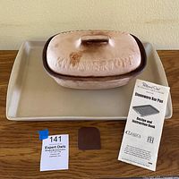 Knuspertopf bread maker with lid, Pampered Chef stoneware bar pan, silicone holder piece and instruction booklet displayed on a table.