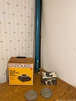 Overall display of Kodak Carousel 600H projector box, foldable projection screen in blue roller case, Premier Photowipes box, and two metal film cannisters on corner shelf.